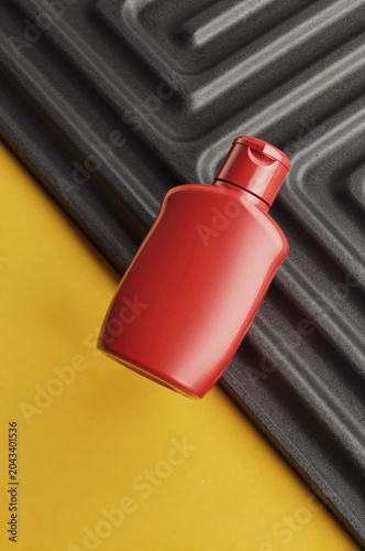 Red plastic bottle rests diagonally across a textured dark grey tray and a solid yellow surface. Harsh light casts sharp shadows against the corrugated ridge patterns of the matte base.