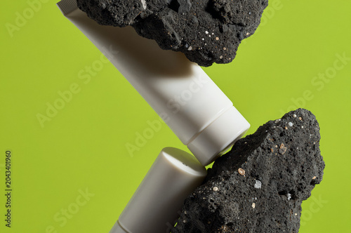 Cosmetic tube rests diagonally between two dark volcanic rocks against a lime green backdrop. Hard shadows cast by the overhead light emphasize the gritty stone and smooth plastic surface.