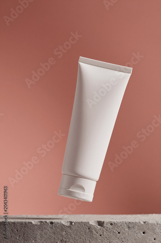 Cosmetic tube floats above a rough stone surface against a terracotta backdrop. Soft shadows define the matte white plastic while even light illuminates its smooth form.