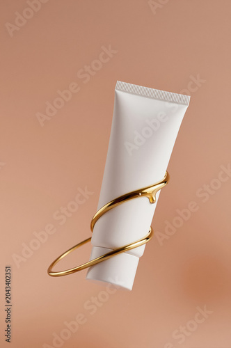 Cosmetic tube rests diagonally against a beige backdrop while golden metal rings wrap around its middle. Soft side lighting creates gentle shadows on the matte white surface.