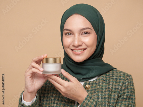 Smiling young Asian Muslim woman wearing green hijab holding skincare cream jar with gold lid against beige background beauty product portrait