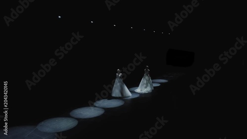 Wide aerial shot of two elegant models in white wedding gowns walking along dark runway illuminated by circular LED ring lights at bridal fashion show. Perfect for wedding, fashion, and luxury content