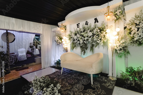 Foto Elegant romantic wedding stage featuring a luxurious white curved chaise lounge in front of a stunning white floral backdrop with E & A initials, chandeliers, and lush greenery