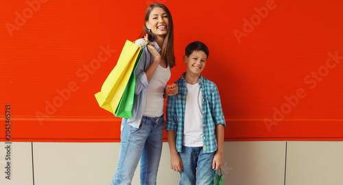 Happy smiling mother and son child together with shopping bags in city on red wall background