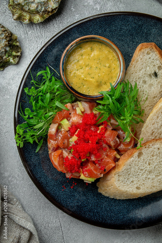 Top view of salmon tartare with avocado, red caviar, bread slices, fresh herbs and creamy sauce on a dark plate