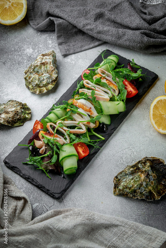 Top view seafood salad with squid, cucumber ribbons, tomatoes, olives and fresh greens served on black slate with lemon