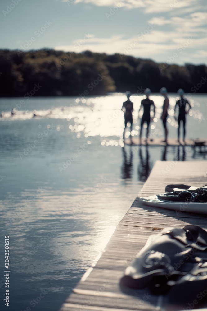 Obraz premium Triathletes standing on wooden jetty by lake before swimming competition. Silhouette of man and woman swimmer preparing for race. Outdoor sports training and healthy lifestyle concept.