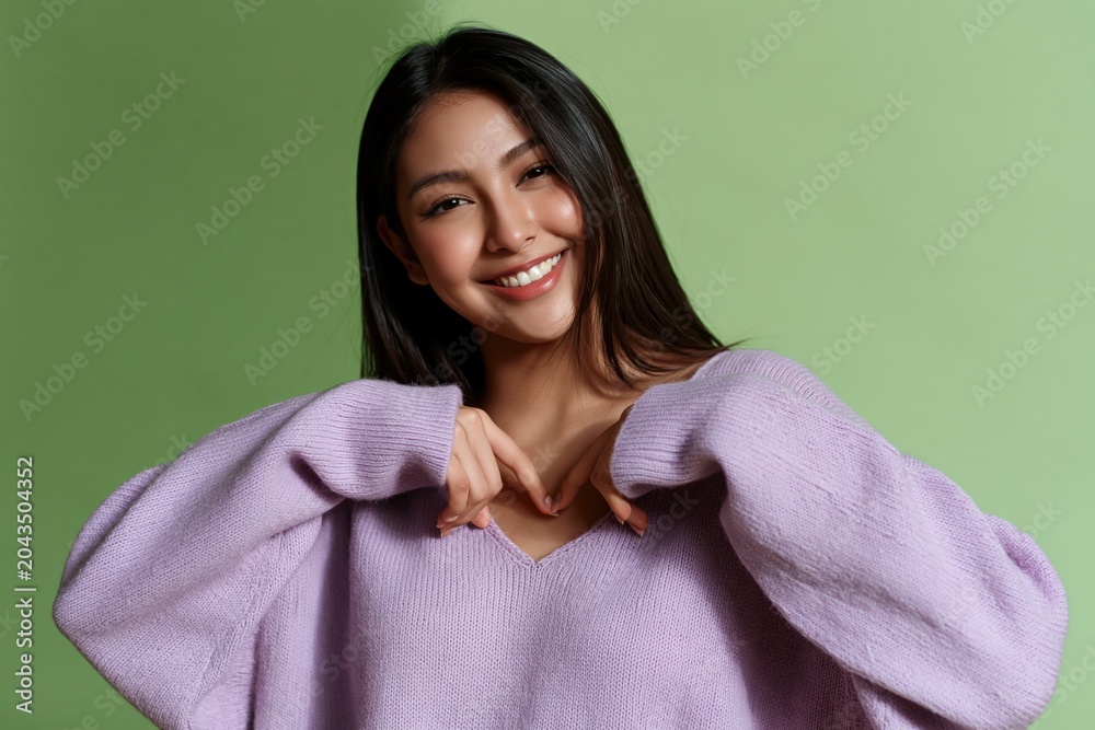 Fototapeta premium Happy woman forming a heart shape with hands in oversized lilac sweater on green backdrop for social media content and banner design