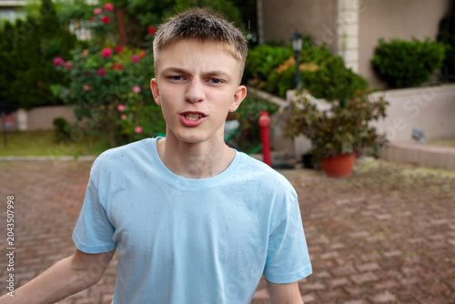 Angry teenage boy with furrowed brows stands in garden courtyard, speaking emotionally. Red fire hydrant and roses in background