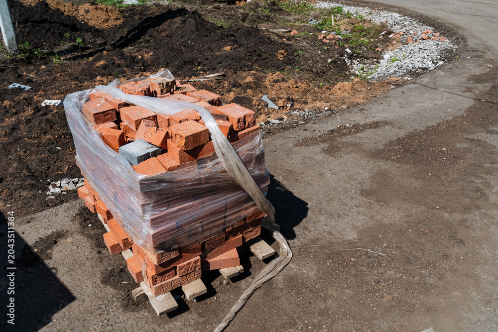 Fototapeta premium Pallet Of Bricks Roadside Assembly. Urban DIY Project With Chipped Brick Units. Masonry Supplies Consisting Of Red Bricks On Pallet With Damaged Wrapping Ready For Building Repairs