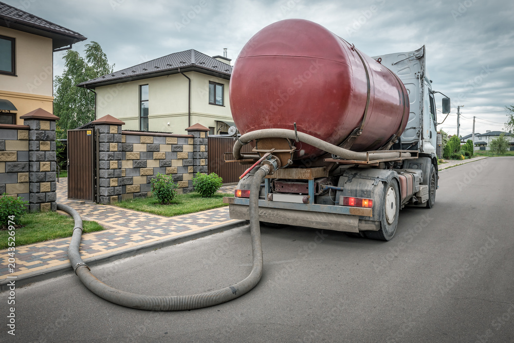 Fototapeta premium Red sewage vacuum truck pumping out a septic tank through a long hose at a residential suburban house.