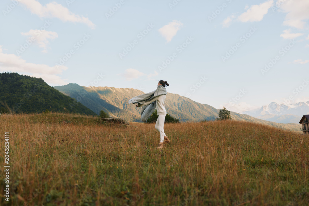 Fototapeta premium Young woman in casual clothes enjoys freedom and fresh air running through grassy field with mountains in background under blue sky with clouds.