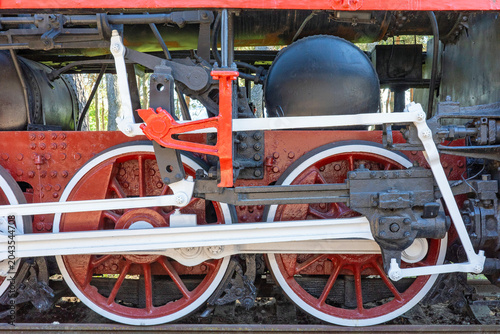 A detailed close-up view capturing the complex mechanical assembly of a historic steam locomotive, showcasing its intricate iron drive wheels and red-and-white painted rods.