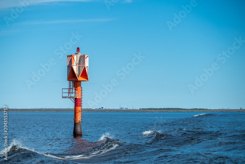 Lighthouse. Marine navigation buoy in the sea waves view from a moving ferry boat, coastal shipping lane light beacon with splashing water in the foreground and shoreline on the horizon.