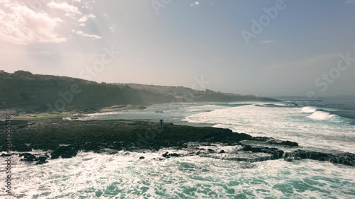 Aerial drone view with at the camera passing in and passing by a fisherman on the rocks at the beach.
