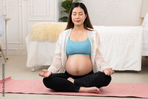 A pregnant woman practices mindfulness meditation on a pink yoga mat in a serene bedroom. With eyes closed and hands resting on knees, the scene highlights prenatal yoga, relaxation