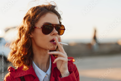 Foto Cheerful woman outdoors, sunglasses on, red jacket, smoking cigarette, sunlit mo