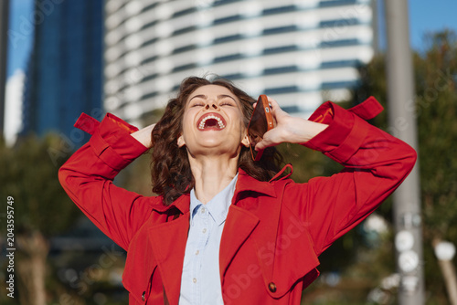 Tablou pe pânză Young woman in a red coat laughs joyfully while holding a phone to her ear outdoors in an urban environment on a sunny day
