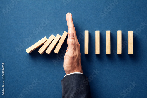 Businessman hand stops falling wooden dominoes on a dark blue surface to demonstrate the prevention of a major risk and the importance of professional intervention for maintaining operational.
