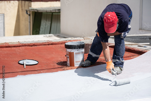 Worker applying white waterproofing liquid membrane on a red roof with a paint roller