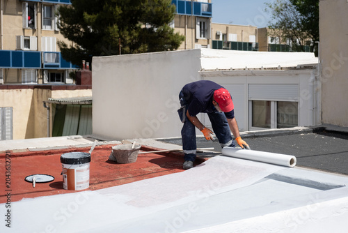 Construction worker applying a white waterproof membrane on a red flat roof during renovation