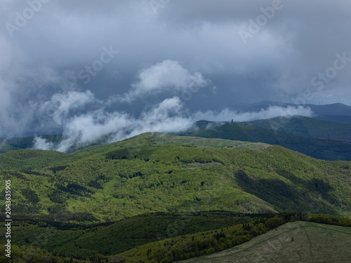 Rolling hills covered in vibrant green trees are shrouded in low-lying mist under a dramatic, overcast sky with scattered clouds