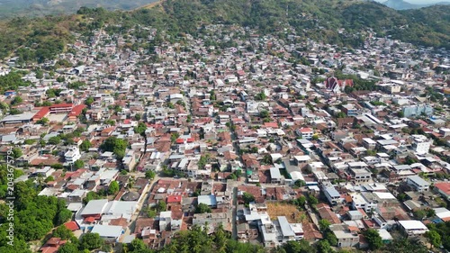 Aerial drone view captures the vast urban sprawl of Tecpan de Galeana Guerrero, Mexico. Houses densely packed with lush green hills and a tranquil river completing the scenic landscape.