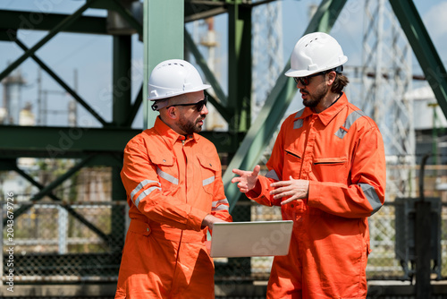 Two male refinery engineers discussing maintenance plan with laptop at petrochemical site, coordinating pipeline safety inspection, compliance audit, field operation workflow and hazard review