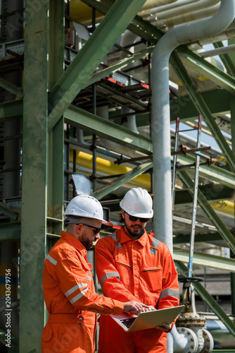 Male refinery technician team checks laptop under pipeline rack, managing petrochemical maintenance checklist, safety inspection audit, operation monitoring workflow inside industrial plant site