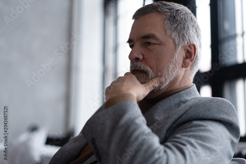 Portrait of handsome bearded mature businessman looking away thoughtfully while sitting on sofa.