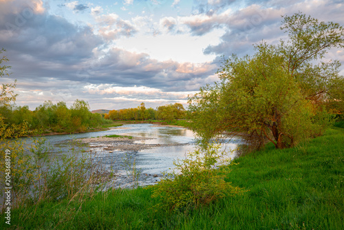 Photography Tranquil landscape of river Mures , central Transylvania, Romania, with green trees and partly cloudy sky at golden hour in spring