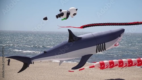 A giant realistic shark kite and a pair of panda kites flying near the water on a sunny beach day with people relaxing below.