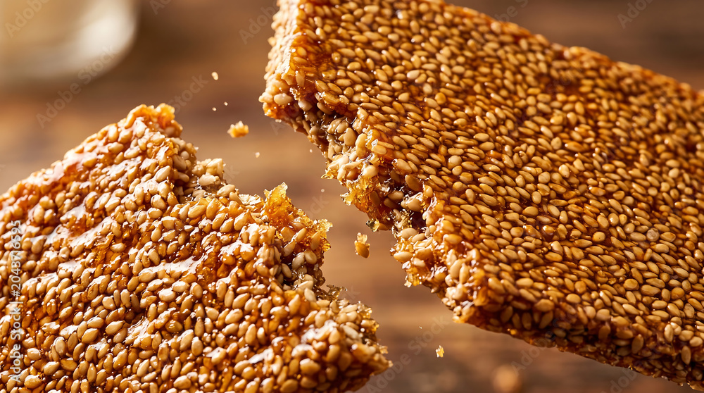 Obraz premium Macro dynamic action shot of a crunchy golden sesame seed bar being snapped in half with tiny flying crumbs over a warm blurred wooden background