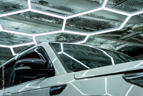 Close view of a white sports car inside a modern indoor setting, with strong reflections from geometric ceiling lights across the body and windshield