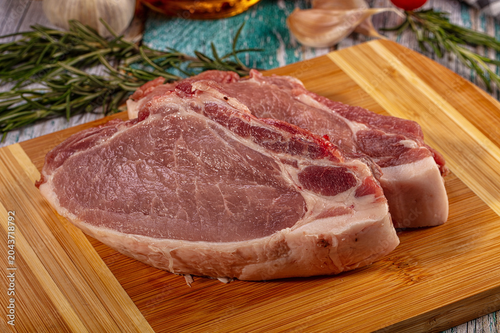 Fototapeta premium Raw pork loin chops on bamboo cutting board with rosemary, garlic, and tomatoes