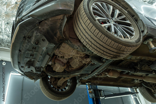 Underside view of a car elevated on a service lift inside a repair workshop, showing wheels, suspension parts, exhaust components, and mechanical underbody