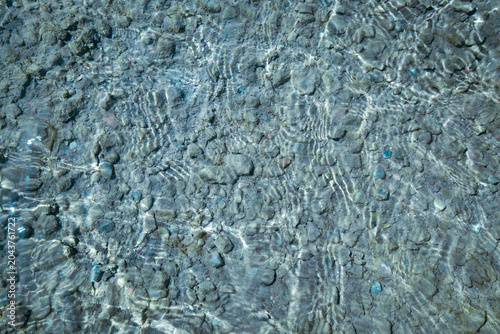 Abstract close-up view of clear shallow water over a rocky seabed, with sunlight reflections and gentle ripples creating natural patterns on stones and sand.