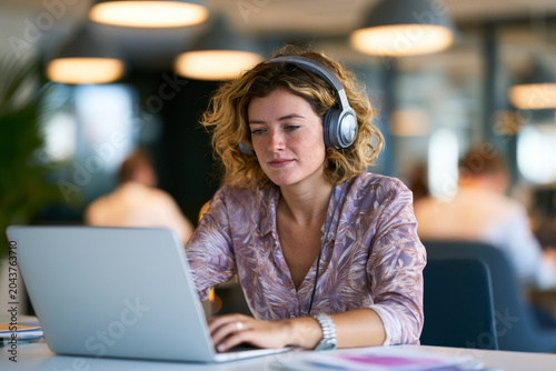 Young woman wearing headphones while working on a laptop in a modern office during a hybrid workday, illustrating focus, productivity, flexible schedules, and contemporary workplace routines