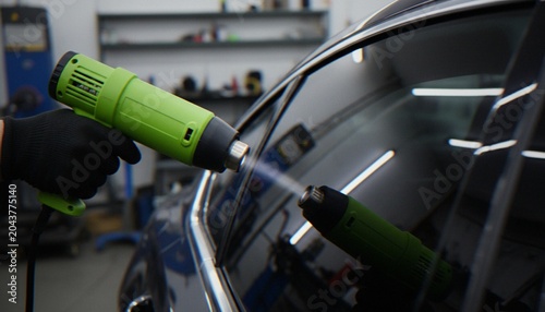 Fotografie A close-up shot of a car detailer using green heat guns to apply protective film on a vehicle window in a professional workshop