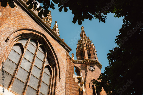 Low-angle view of a Gothic brick church showing a tall ornate spire, clock face, and large arched leaded windows. Warm sunlight highlights the textured red