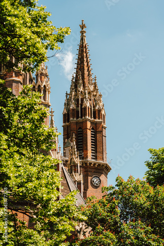 Tall Gothic church tower with a richly detailed brick spire and decorative pinnacles rising above surrounding leafy trees on a clear blue sky day. Visible