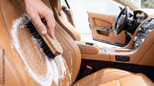 Detailer cleaning perforated leather car seat with brush and foam. Man maintaining luxury vehicle interior. Professional auto detailing service for upholstery maintenance and hygiene.