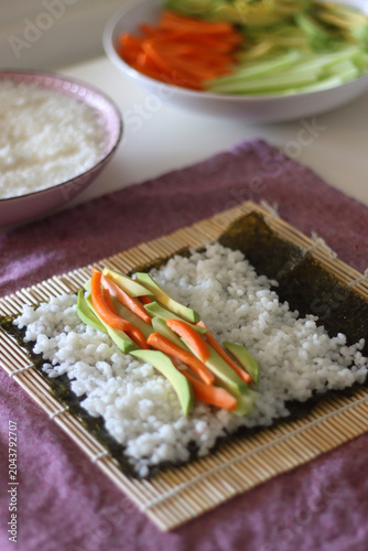 Making vegan sushi rolls at home. Bamboo mat, nori sheets, rice, cut vegetables and soy sauce on the table. Selective focus.