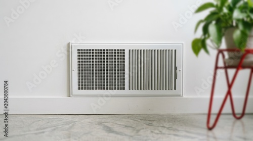 White wall mounted air vent grill on floor with minimalist interior design and small green plant in blurred background.