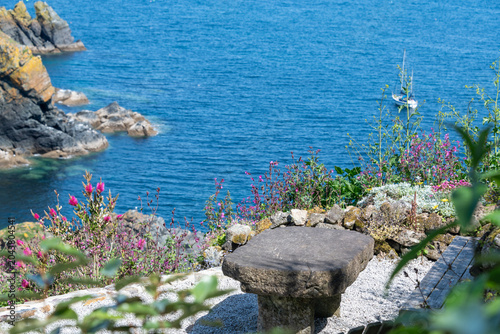 A stone table in a small coastal garden, overlooking the sea and cliffs with summer flowers.