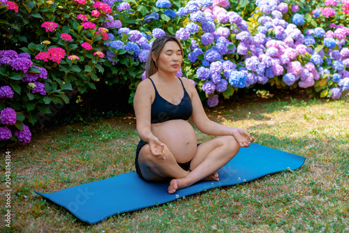 Pregnant woman sitting cross legged on a yoga mat with eyes closed, finding peace and mindfulness through meditation amidst vibrant hydrangeas and natural surroundings