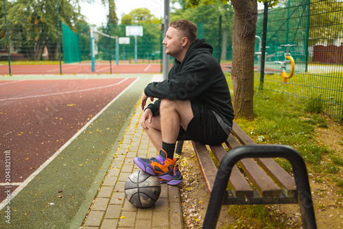Full length profile view of a young athletic man sitting on a wooden bench with a basketball at his feet on an outdoor court