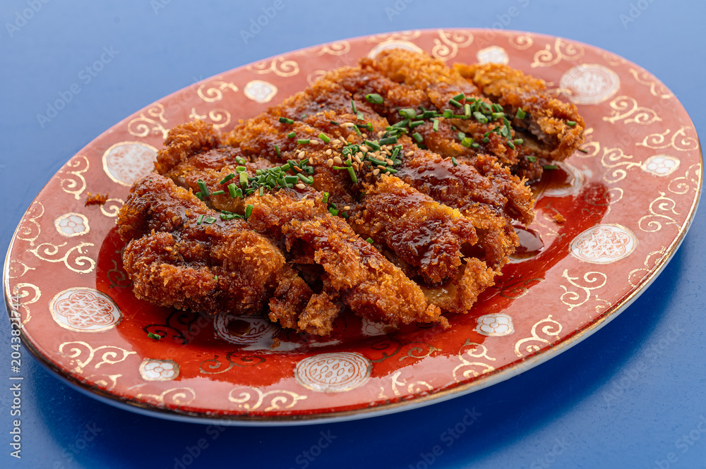Fototapeta premium Chinese-style crispy fried pork coated with savory sauce, topped with sesame seeds and chopped chives, served on a decorative ceramic plate.