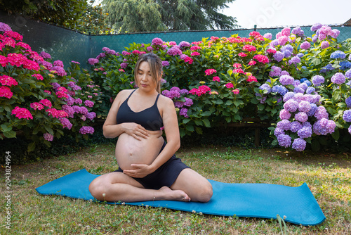 Pregnant woman sitting in lotus position on a yoga mat in a garden, gently touching her belly while finding peace and tranquility surrounded by blooming hydrangeas