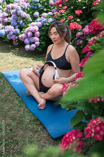 Pregnant woman sitting cross legged on a yoga mat, placing headphones on her belly, closing her eyes in a moment of prenatal meditation and relaxation surrounded by colorful hydrangeas.Music therapy